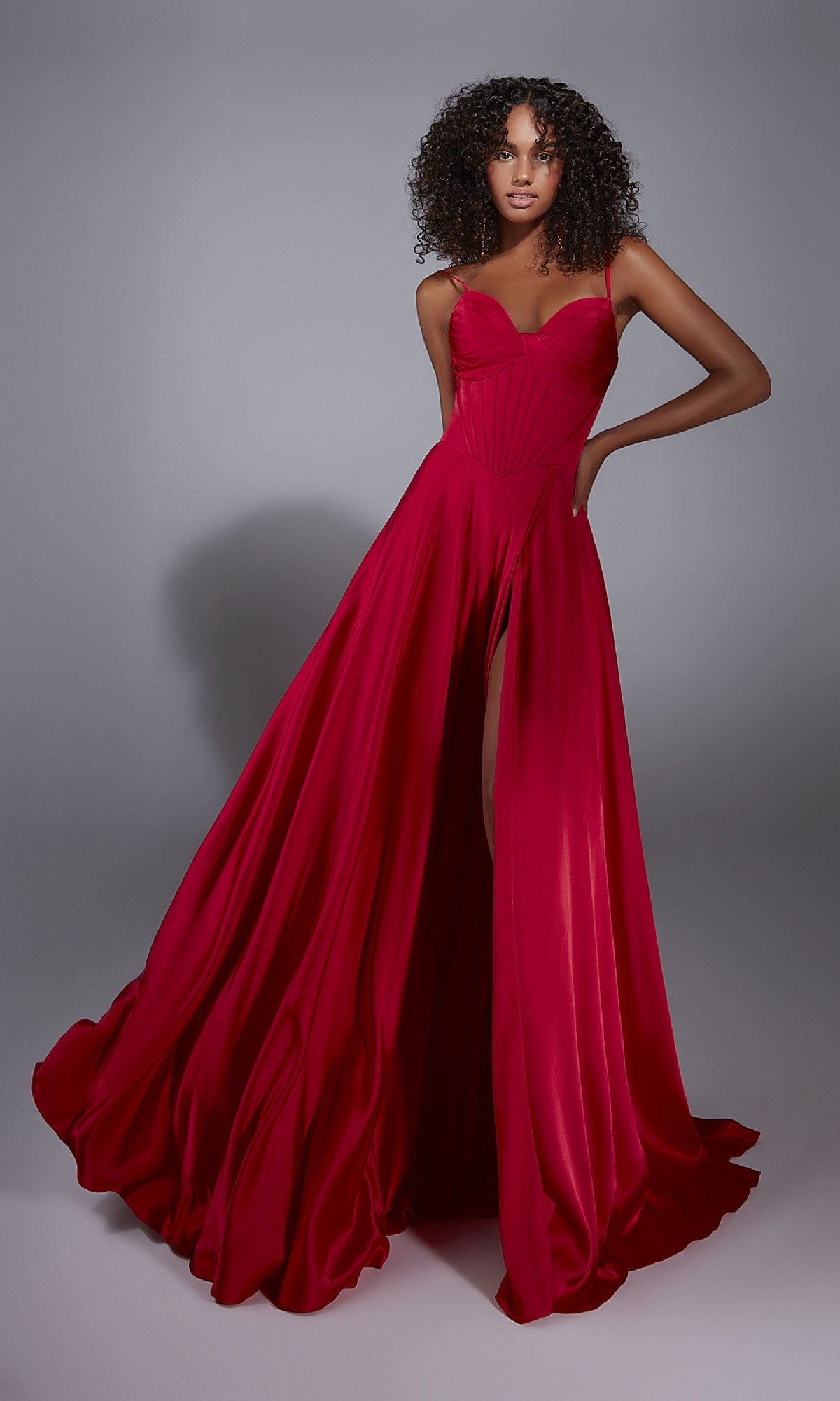 A woman wears the Alyce 61828 long prom dress—a sleeveless, bright red A-line gown with a high leg slit and corset bodice—posing against a plain gray background.